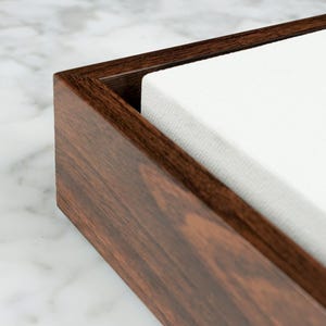 May include: Close-up of a rectangular wooden box with a dark brown finish. The box contains a stack of white fabric. The wood grain is visible, adding texture to the design. The box is likely used for storage or display.