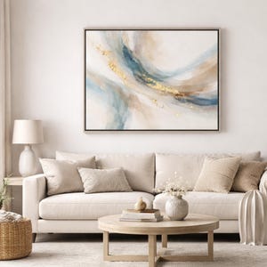 May include: Abstract art print in a wooden frame. The artwork features flowing blue, beige, and gold watercolor-style strokes. The painting hangs above a beige sofa with decorative pillows, a round wooden coffee table, and a white lamp.