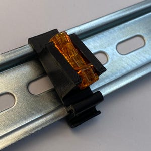 May include: A black plastic electrical component with an orange, translucent center is clipped onto a silver metal rail. The rail has several oval-shaped openings. The component appears to be a connector or terminal block.
