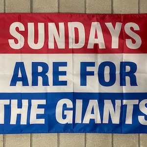 May include: A rectangular flag with a red top, white center, and blue bottom. The flag displays the text "SUNDAYS ARE FOR THE GIANTS" in large, white capital letters. The flag is hanging against a gray brick wall.