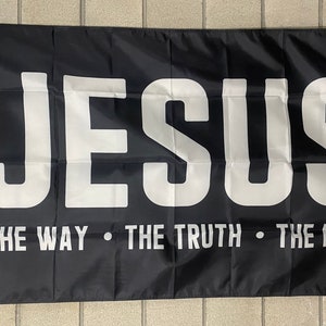 May include: Black flag with the word "JESUS" in large white capital letters. Below, the text reads "THE WAY • THE TRUTH • THE LIFE" in white. The flag has a white border and grommets for hanging.