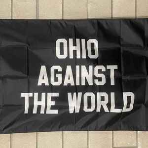 May include: Black flag with white text that reads "OHIO AGAINST THE WORLD". The flag is rectangular and has grommets for hanging. The text is in a bold, sans-serif font.