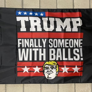 May include: Black and red flag with white text that reads "TRUMP FINALLY SOMEONE WITH BALLS!" and a cartoon image of Donald Trump. There are stars in the top left corner and bottom center of the flag.