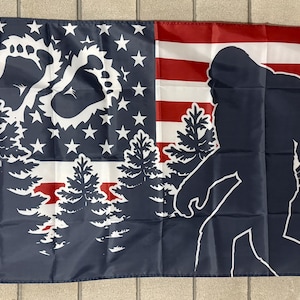 May include: A flag featuring a design that combines the American flag with a Bigfoot silhouette and footprint. The left side has the stars and footprint, with trees. The right side has the Bigfoot silhouette and red and white stripes.