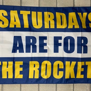 May include: A rectangular flag with the text "SATURDAYS ARE FOR THE ROCKETS" in yellow block letters. The flag has a blue top and bottom border, a white center section, and a metal grommet on the left side.