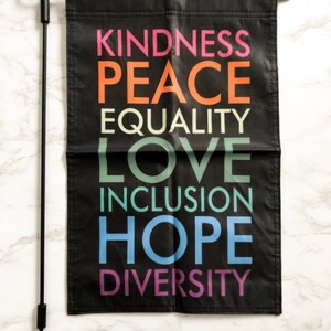 May include: Black garden flag with the words "KINDNESS, PEACE, EQUALITY, LOVE, INCLUSION, HOPE, DIVERSITY" in various colors. The flag is hanging on a black metal pole.