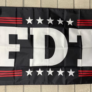 May include: Black flag with the white letters "FDT" in the center. Above and below the letters are five white stars. Red stripes are above and below the stars. The flag is rectangular.