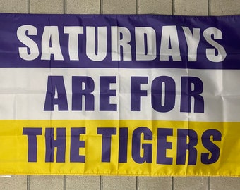 Könnte beinhalten: Rechteckige Flagge mit dem Text "SATURDAYS ARE FOR THE TIGERS" in lila Blockbuchstaben. Die Flagge ist in drei horizontale Abschnitte unterteilt: oben lila, in der Mitte weiß und unten gelb. Die Flagge hat Ösen zum Aufhängen.