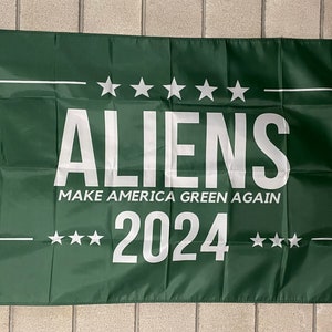 May include: A green flag with white text that reads "ALIENS MAKE AMERICA GREEN AGAIN 2024" with five white stars above and below the text.