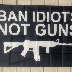 May include: Black flag featuring white text and a white rifle silhouette. The text states "BAN IDIOTS NOT GUNS". The flag is rectangular with a white border on the left.