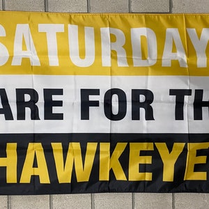 May include: A rectangular flag with horizontal stripes of gold, white, and black. The text "SATURDAYS ARE FOR THE HAWKEYES" is printed in bold, black and gold letters. The flag is hanging against a light gray brick wall.
