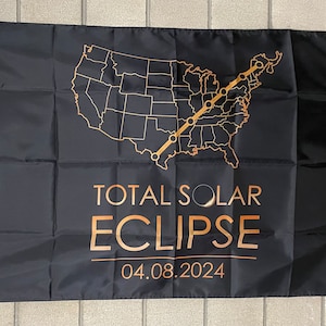 May include: Black flag with a gold outline of the United States map and a path of the total solar eclipse across the country. The text "TOTAL SOLAR ECLIPSE" and the date "04.08.2024" are printed in gold.