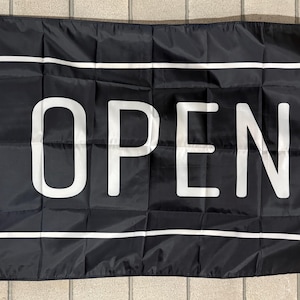 May include: A rectangular black flag featuring a white rectangular border and the word OPEN printed in large white capital letters in the center. The flag has a white vertical strip on the left side with two metal grommets for mounting purposes.