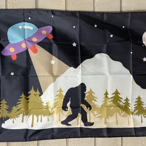 May include: Black flag featuring a cartoon scene of a UFO beaming a light onto a Bigfoot silhouette in a forest. The background includes a full moon and stars. The UFO is purple, blue, and red.