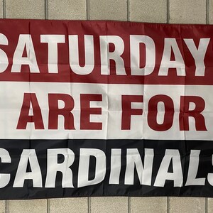 May include: A rectangular flag with the words "SATURDAYS ARE FOR CARDINALS" in large white letters. The flag is divided into three horizontal sections: maroon at the top, white in the middle, and black at the bottom. The flag has a gold grommet.