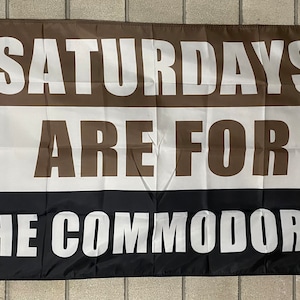 May include: A rectangular flag with horizontal stripes of brown, white, and black. The words "SATURDAYS ARE FOR THE COMMODORES" are printed in large, white capital letters across the flag. The flag has grommets for hanging.