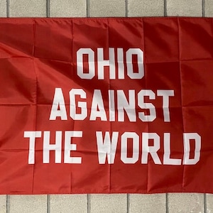 May include: A red flag with white text that reads "OHIO AGAINST THE WORLD". The flag is rectangular and has a white border on the left side with grommets for hanging. The text is in a bold, sans-serif font.