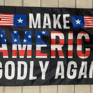 May include: A black flag with the text "MAKE AMERICA GODLY AGAIN" in white letters. The text is surrounded by red, white, and blue stars and stripes.