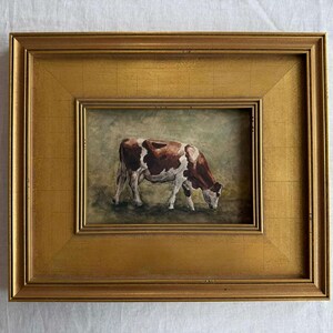 May include: A framed painting of a cow grazing in a field. The cow is brown and white, and the painting is set in a gold-colored frame. The artwork is a landscape painting.