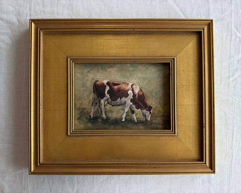 May include: A framed painting of a cow grazing in a field. The cow is brown and white, and the painting is set in a gold-colored frame. The artwork is a landscape painting.