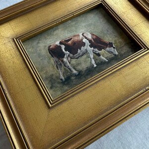 May include: A framed painting of a cow in a field. The cow is brown and white, and the frame is gold. The painting is set in a gold frame with a double border.