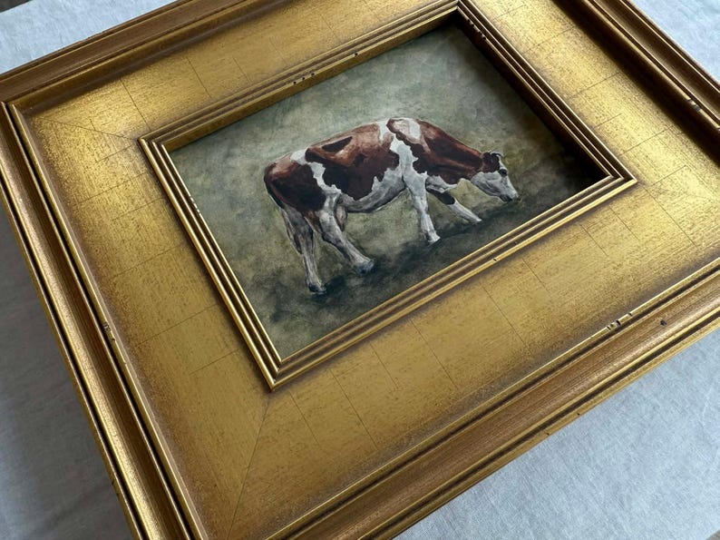 May include: A framed painting of a cow in a field. The cow is brown and white, and the frame is gold. The painting is set in a gold frame with a double border.