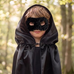 Könnte beinhalten: Ein Kind trägt einen schwarzen Satin-Kapuzenmantel und eine flauschige schwarze Maske. Der Umhang wird mit einem dekorativen Verschluss befestigt. Der Hintergrund ist eine verschwommene Waldszene.