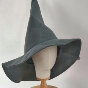 Peut inclure: Un chapeau de sorcière gris foncé avec un large bord et une couronne pointue. Le chapeau est présenté sur une tête de mannequin et un support en bois, sur fond blanc. Le tissu du chapeau semble être du feutre doux.