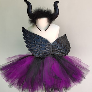 Peut inclure: Ensemble de costume avec un serre-tête à cornes noires, des ailes noires et une jupe tutu superposée noire et violette. Le costume est présenté sur un mannequin blanc.