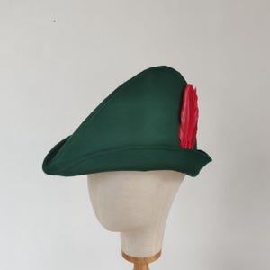 May include: A dark green hat with a red feather attached. The hat is displayed on a mannequin head. The hat has a curved brim and a pointed top. The background is a plain white wall.