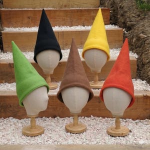 Peut inclure: Cinq chapeaux de gnome colorés en vert, noir, marron, jaune et orange sont exposés sur des têtes de mannequin. Les chapeaux ont une forme pointue classique et sont faits d'une matière douce. Les chapeaux sont sur des socles en bois.
