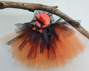 Puede incluir: Un tutú y un sombrero de bruja con temática de Halloween. El tutú está hecho con tul naranja y negro en capas, adornado con lentejuelas plateadas. El sombrero es naranja con un diseño de telaraña negra y una cinta negra. El conjunto está suspendido de una rama de madera.