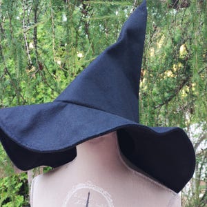 May include: A dark navy witch's hat with a wide, undulating brim and a tall, pointed crown. The hat is made of a dark material. The hat is displayed on a mannequin with an Eiffel Tower design.
