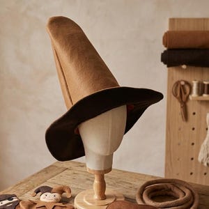 Op de afbeelding: Een bruine heksenhoed met een brede rand wordt getoond op een mannequinhoofd. De hoed is gemaakt van vilt en heeft een hoge, puntige kroon. Andere kostuumaccessoires, waaronder een masker en poppen, liggen op een houten tafel.