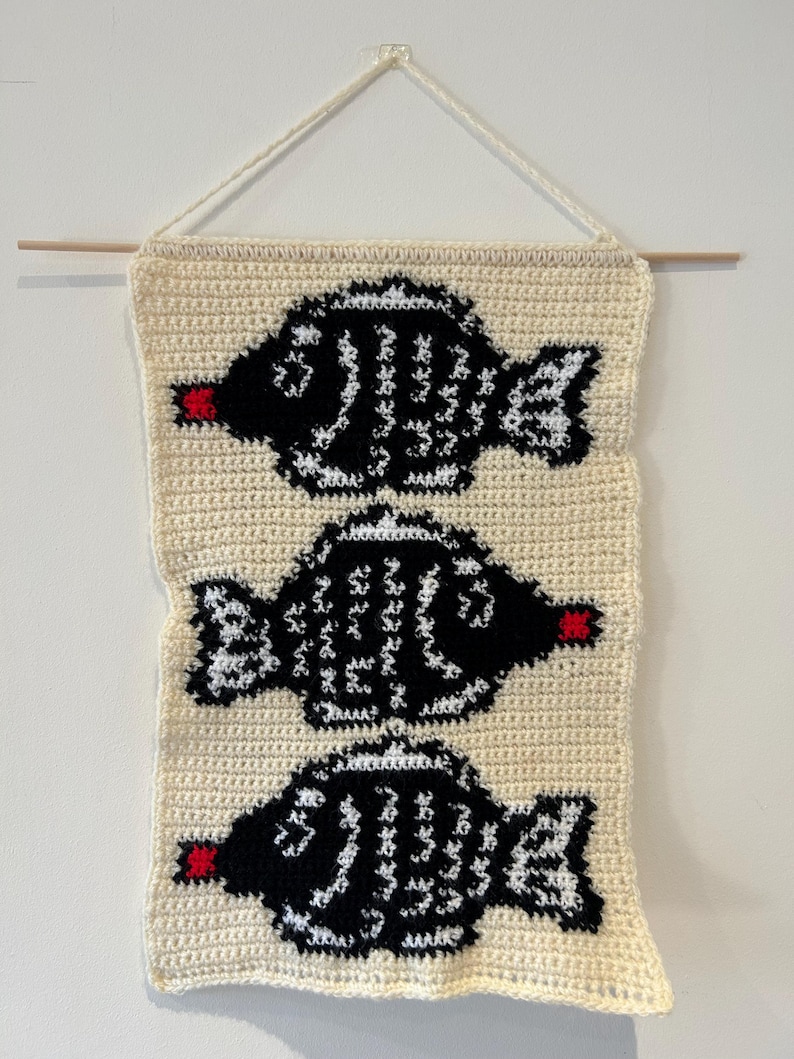 May include: A crocheted wall hanging featuring three black and white fish with red mouths. The fish are arranged vertically on a cream-colored background.