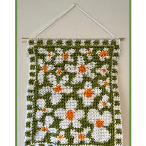 Puede incluir: Un tapiz de pared tejido a crochet con un patr&oacute;n de margarita en blanco, verde y naranja. El tapiz est&aacute; colgado en una pared blanca con una varilla de madera.