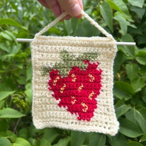 May include: A cream-colored, crocheted wall hanging featuring a red strawberry design with green leaves. The strawberry has small yellow accents. The square banner is held by a cream-colored loop and a white dowel rod.