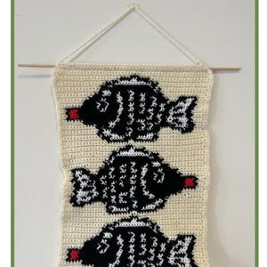May include: A crocheted tapestry featuring three black and white fish with red mouths. The tapestry is hanging on a white wall with a wooden dowel rod. The text "SOY SAUCE FISH CROCHET TAPESTRY" is at the top of the image. The text "By jf00creates" is at the bottom of the image.