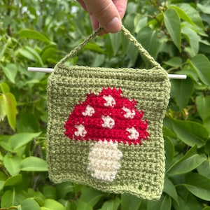 May include: A small, square, crocheted wall hanging featuring a red mushroom with white spots on a green background. The mushroom is centered on the square, with a white stem. The hanging has a green border and a loop for hanging.