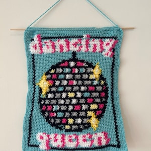 Może przedstawiać: Wiszący dywanik wykonany na szydełku z niebieskim tłem i wzorem czarnej, białej i różowej kuli dyskotekowej. Tekst "Dancing Queen" jest napisany różowymi i białymi literami.