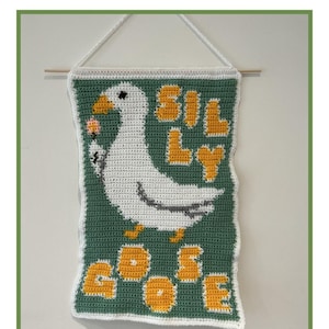 May include: A crochet tapestry wall hanging with a green background, a white goose holding a pink flower, and the words "Silly Goose" in yellow letters.