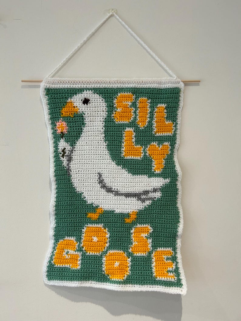 May include: A crocheted wall hanging featuring a white goose with a pink ice cream cone, against a green background with the words "Silly Goose" in yellow letters.