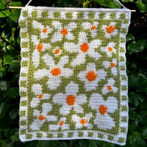 Puede incluir: Un tapiz de pared tejido a crochet con un patr&oacute;n geom&eacute;trico verde y blanco con margaritas blancas con centros naranjas.