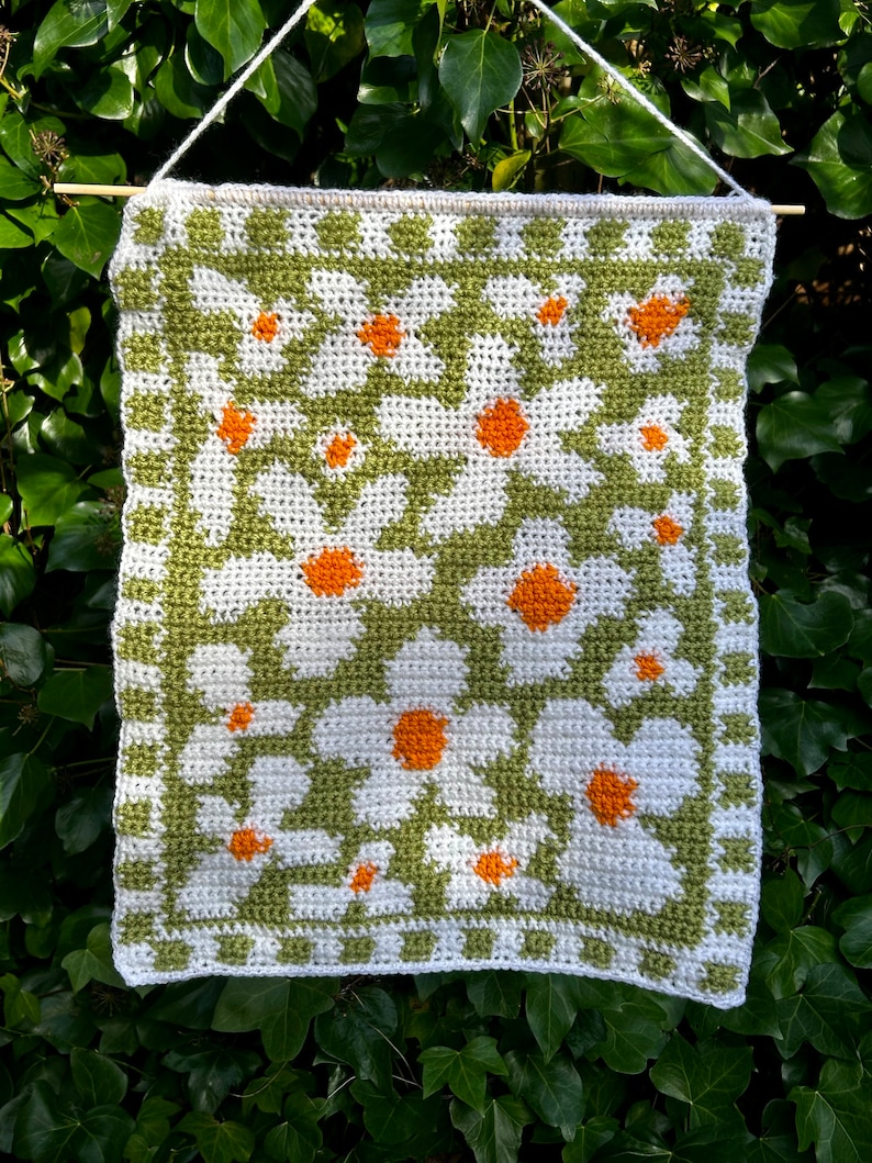 Puede incluir: Un tapiz de pared tejido a crochet con un patr&oacute;n geom&eacute;trico verde y blanco con margaritas blancas con centros naranjas.