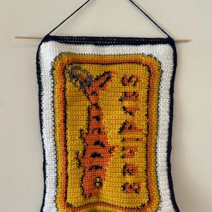 May include: Handmade crochet wall hanging featuring a yellow and orange design with the word "TEXAS" in black. The banner has a white border with a navy blue trim and is suspended by a wooden dowel and black string.