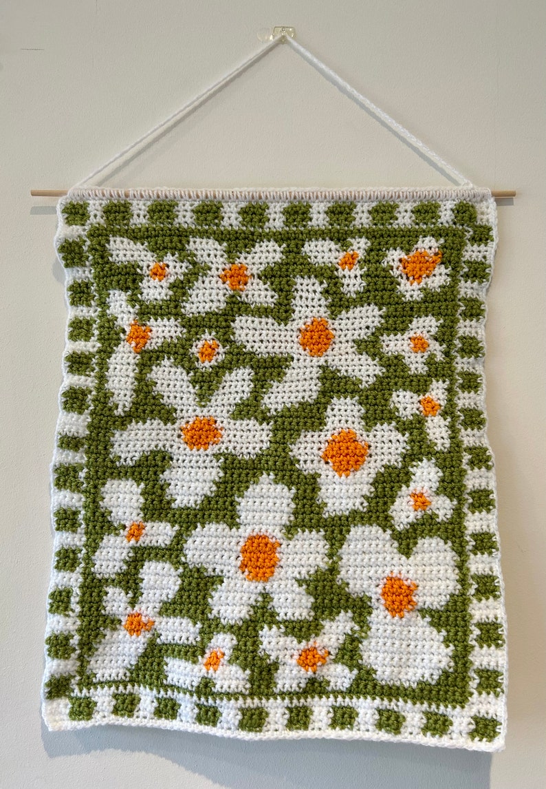 Puede incluir: Un tapiz de pared tejido a crochet con un patr&oacute;n de margaritas verdes y blancas. Las margaritas tienen centros naranjas.