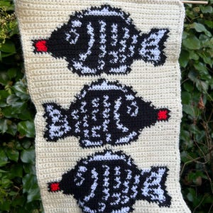 May include: A crocheted wall hanging featuring three black and white fish with red accents. The fish are arranged vertically on a cream-colored background.
