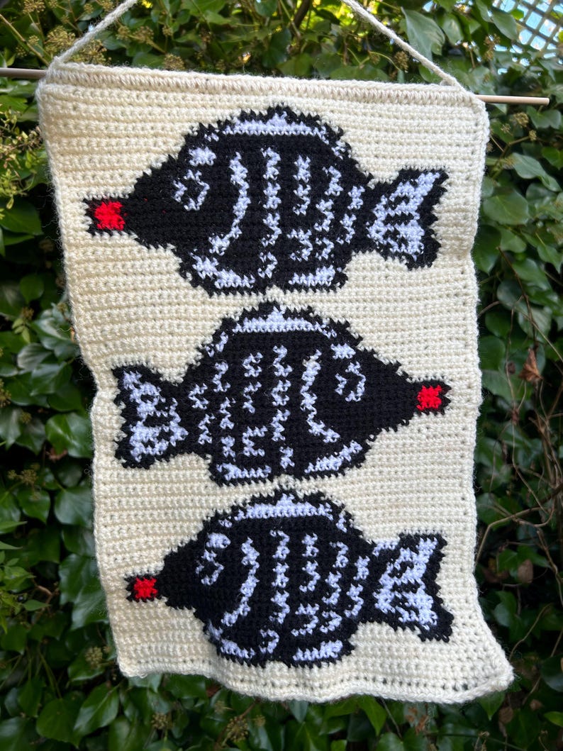 May include: A crocheted wall hanging featuring three black and white fish with red accents. The fish are arranged vertically on a cream-colored background.
