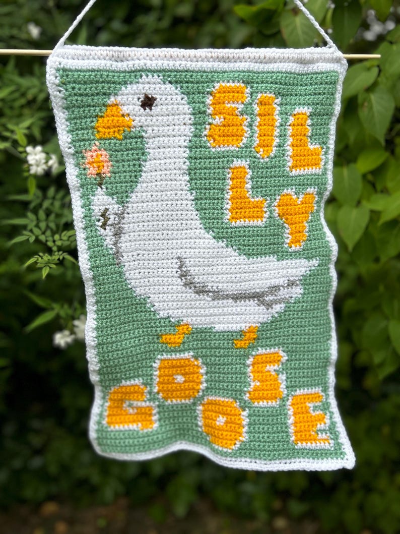 May include: A crocheted wall hanging featuring a white goose with a flower in its beak. The words "Silly Goose" are written in yellow yarn on a green background.