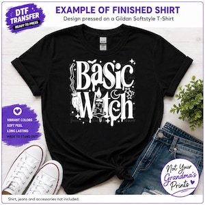 May include: A black short-sleeved t-shirt featuring a white graphic print with the text Basic Witch, a broomstick, a candle, a crescent moon, stars, and a bat. The shirt has a crew neckline and is displayed with white sneakers and blue jeans.